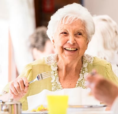 A smiling assisted living resident is enjoying a meal with friends in a well-lit dining hall.